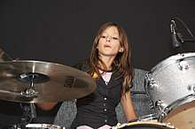 A teenager is seated behind a drum kit. One arm is obscured by the cymbal stand at her right. She is looking in that direction. Her other hand is down her left side. A drum to her left is miked.