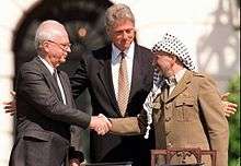A man in a dark suit on the left shakes the hand of a man in traditional Arab headdress on the right. Another man stands with open arms in the centre behind them.