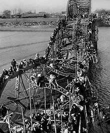 Black-and-white photo of people crossing a river via a destroyed bridge