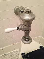 A scuffed and oxidized metal flushometer valve with a porcelain handle attached to the back of a toilet