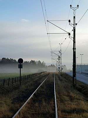 A railway line with overhead wires, supported by latticework gantries on its right, seen just from its side. On the left is the rear of a sign with a circular piece above an equally-sized rectangular one. Golden-colored grasses rise from in between the ties, and there is a fogged area of woodland around the tracks in the distance under clouds with blue sky above them. On the right is a road; a fence separates the track right-of-way from a green field on the left.