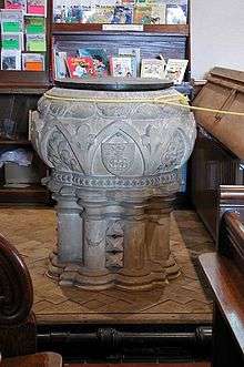 A wide, round-bodied font of pale stone with several rounded pillars forming its base.  The stonework is elaborately carved.  Around the top is a thin yellow rope, and a bookshelf stands in the background.