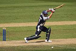 A batsman playing a square drive. He is wearing a blue cricket helmet, white gloves, blue pads, a blue-white top and bottom. He is holding a cricket bat.