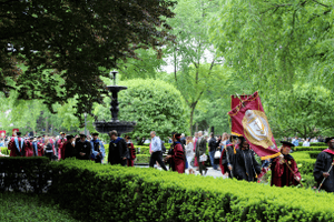 Students and faculty in commencement garb walking down pathway among greenery.