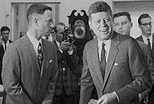 Black-and-white film screenshot showing the main character on the left looking towards another man, President Kennedy, (voiced by actor Jed Gillin), on the right. Kennedy is smiling and looking to his left. In the background, several men are looking in different directions and one is aiming a camera.