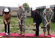 A woman, two army soldiers wearing Army Combat Uniforms, and one man in a suit, all wearing hardhats carrying shovels with freshly dug dirt.