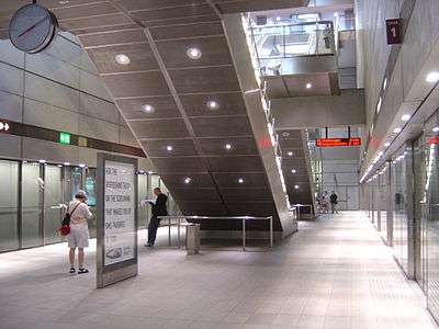 A square steel and glass room with some people waiting. In the middle are the backs of two escalators.