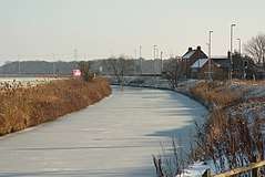 The Fosdyke curves through the picture, its surface smooth white ice.  The two banks are brown with dead grass, and the fields either side crop-less and white with hoar. Farther along the bank, in the middle distance, a small group of redbrick buildings are grouped together.  Lamp posts are ranged on the right bank, facing away from the canal. They are for lighting the adjacent, but invisible, A57 road.