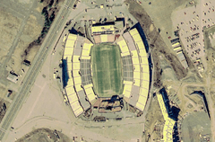 An aerial view of a large sports stadium with a four-lane road next to it on the left and the beginnings of a similar structure at lower right