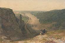 Two people on a cliff top look along the length of a river with wide muddy banks which snakes through a gorge towards the distant city of Bristol. A line of small boats sail along the river.