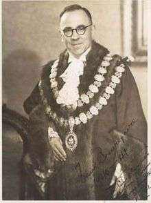 Middle aged man with receding dark hair, wearing circular glasses. He is wearing a dark robe with two chains of circular decorative disks, a fluffy light-coloured tie, and is standing next to a ceremonial chair. The subject of the photograph has signed it.