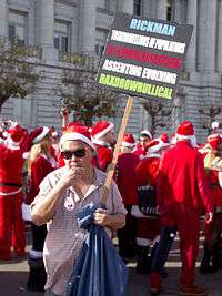 Frank Chu, wearing a Santa hat, holds a sign that reads "Rickman / Toskrojrellions of populations / CBS: Nesprofrolluxul rocketcenters / Assenting evolving / Raxdrowrullical"