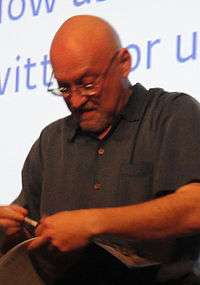 A caucasian, bald man wearing glasses and a dark shirt. He looks down at a book and pen held in his hands.