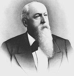 Formal portrait, head and shoulders, of serious-looking man of about 60 dressed in a dark suit jacket and white shirt. He is bald in front, and his remaining hair is white. He has a long, carefully combed white beard.