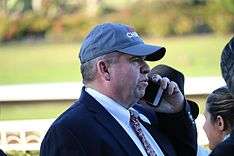 a middle aged man in a dark suit and ball cap, talking on a cell phone
