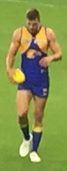 A light-skinned man with brown hair in a blue and gold West Coast Eagles guernsey holding a football in his right hand