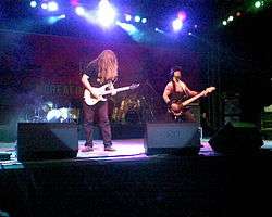 A medium sized concert stage lit in many colors. In the left stands a longhaired guitarist on the right a bass player wearing an armystyle west and helmet. In the background are two drumkits with a drummer.