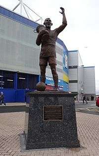 A statute of a man holding a trophy and lifting his left arm into thr air. The statute is in front of a football stadium