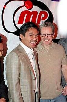 A man with dark hair, wearing a beige suit and white shirt with open collar, stands on the left next to a more lightly complected man with spectacles, stubble and slightly graying hair, wearing a similarly-colored beige shirt with open collar, under a red, white and black banner that says "MP" in curvy stylized lettering. Both are smiling.