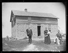 Three adults and two small children in front of two-story house: lower story sod-walled, upper story with clapboard siding
