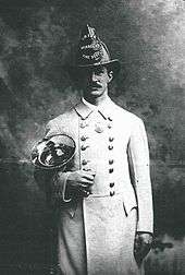 A man in a white coat and fire helmet holding a brass horn