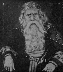 Portrait of a seated man dressed as King Lear. He has long white beard and wears a large crown.