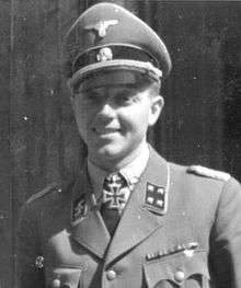 A smiling man wearing a military uniform, peaked cap and a neck order in the shape of a cross. His cap has an emblem in shape of a human skull and crossed bones.