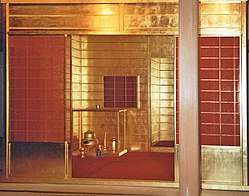 A photograph of an open entrance to a room with gold leaf covering the exterior and interior, there is a red covering on the floor, and several golden utensils and vessels in one corner