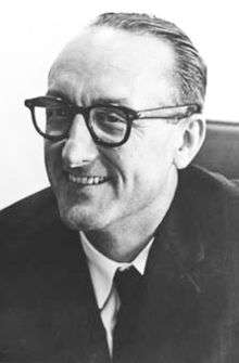 black-and-white image of George Mueller, smiling to the viewer's left in a light shirt, dark tie and suit coat, and wearing dark plastic eyeglasses