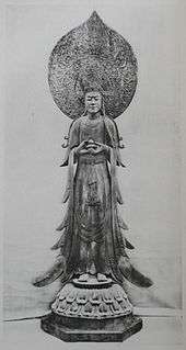 Front view of a standing statue with flowing robes holding a small container in front of her body. There is a large halo behind the head of the statue.