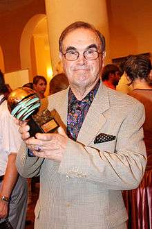 An elderly man with glasses and parting smiling to the camera. He holds a prize in his hands.
