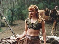 A young blonde woman wearing a green woven tank-top and leather skirt. She holds a wooden fighting staff, a horse can be seen in the background.