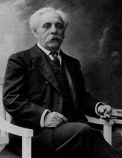 Elderly man with white hair and large white moustache. He is seated, with a cigarette in his left hand