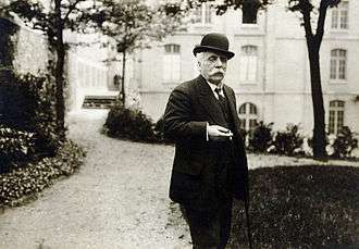 elderly man in a garden. He has a large white moustache, is holding a lighted cigarette, and is wearing a bowler hat