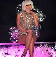 A blond woman in a bob-cut, sitting cross-legged on a transparent platform which is full of bubbles and lit from inside in pink. The woman is wearing a dress made of transparent bubbles of varying sizes. She is holding a microphone in her left hand and appears to be smiling.