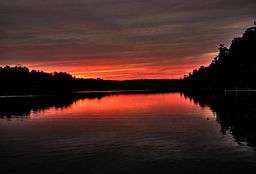 Red, yellow, and purple clouds are reflected in a lake surrounded by silhouetted trees.