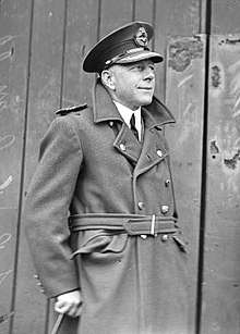 Three-quarter informal portrait of man in military overcoat and peaked cap, holding a cane
