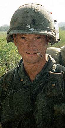 A man is at the center of the image looking at the camera. He is dressed in Vietnam War-era military attire