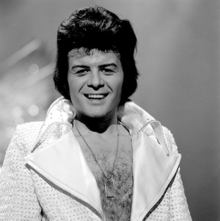 A black-haired man with a hairy chest, wearing a shiny jacket open to the waist, with large lapels, smiles towards the camera.