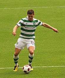 A colour photograph of a footballer, wearing a green-and-white hooped shirt and socks with white shorts and yellow boots. He is controlling the football and preparing to take a shot.