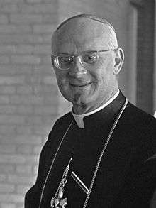 Black and white photograph of a middle-aged man wearing glasses and facing the camera. The photograph is cropped at the chest and the man is wearing a white clerical collar beneath a black cassock with a pectoral cross.