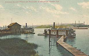 Two steamboats in a dock, with a wooden pier and one gray building. A third vessel is visible behind.