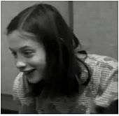 A black and white frame taken from a video of Genie, who is enthusiastically smiling, taken from several feet away. It shows her from the chest up, and Genie is facing slightly to the right of the camera.