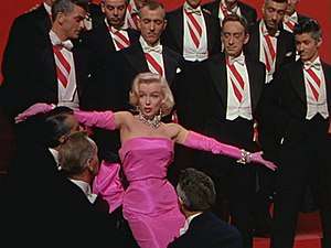 Monroe in Gentlemen Prefer Blondes. She is wearing a shocking pink dress with matching gloves and diamond jewellery, and is surrounded by men in tuxedos