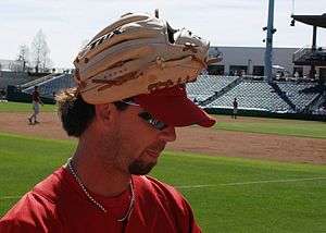 A brown-haired young man with a goatee wearing a tan baseball glove on his hand atop a red baseball cap