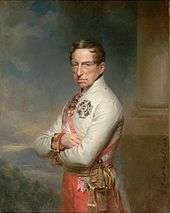 Half-length oil portrait of the Archduke Charles by Georg Decker. Charles wears a white high-collared military jacket of the Austrian army and has a red and white sash over his right shoulder. He wears two decorations, a cross on his breast and another medal at his neck. He has a long fleshy face, short brown hair, and light eyes, and gazes calmly towards the viewer. His arms are folded across his chest.