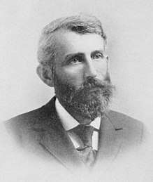 A man in his forties, facing right, with thick black hair, a beard, and a mustache. He is wearing a white shirt, black tie, and black jacket