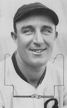 A close-up of a smiling man wearing a light-colored baseball jersey and a dark cap looking into the camera.