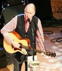 A balding man in a check shirt and dark vest playing a guitar and singing into a mirophone