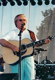 A blond-haired man wearing a white shirt and light blue pants, playing a guitar and singing into a microphone